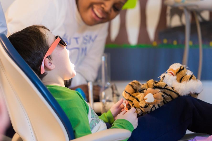 A child, wearing sunglasses and holding a tiger stuffed animal, sitting on dentist chair opening his mouth wide