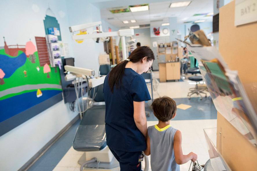 A woman in navy scrubs is walking a male child into a pediatric dental room