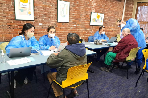 Dental students wearing gowns and masks sit across from members of the Boston Living Center; one student is examining a patients&#039;s mouth.