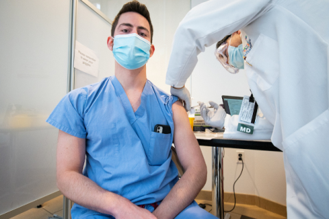 A male medical student in scrubs receives a COVID-19 vaccine in a hospital office. Third- and fourth-year Tufts medical students and dental students in patient-care settings start to receive vaccination shots at Tufts Medical Center
