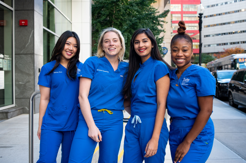Four women in blue scrubs stand outside the School of Dental Medicine. 