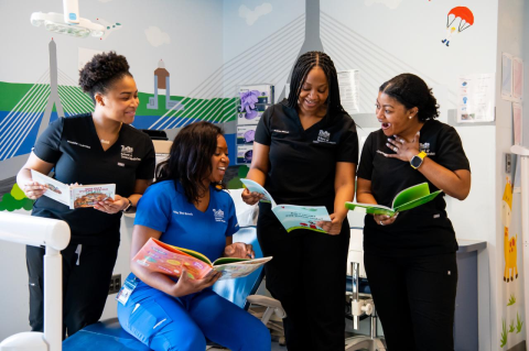 Nickeisha Louis-Elias, D23, Arika Neal-Branch, D24, Ashley Makala, D23, and Amber Courtney, D23, in dental scrubs, look at children&#039;s books while gathered around a dental chair.