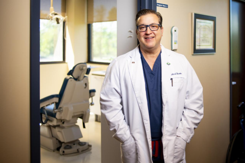 Richard Rosato in blue scrubs and a white dental coat in his office, a dental chair visible in background