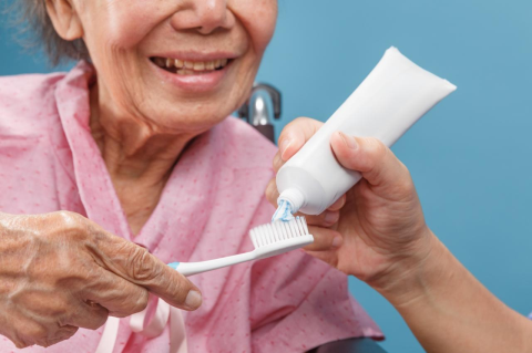 An elderly woman smiling and holding a toothbrush while someone else applies toothpaste
